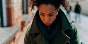 Person wearing a dark green coat and scarf walking outdoors, with focus on upper body and blurred city background