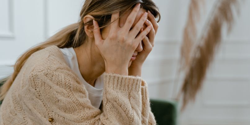 Woman in a beige cable-knit sweater sitting indoors, holding her head in her hand and appearing distressed or contemplative.Woman wearing a beige cable-knit sweater sitting indoors, resting her head on her hand and appearing concerned or thoughtful.